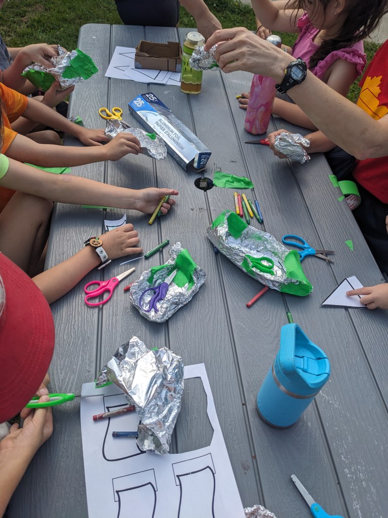 Students sitting at a bench outside, hands-on, folding and shaping pieces of tin foil to construct small boats.