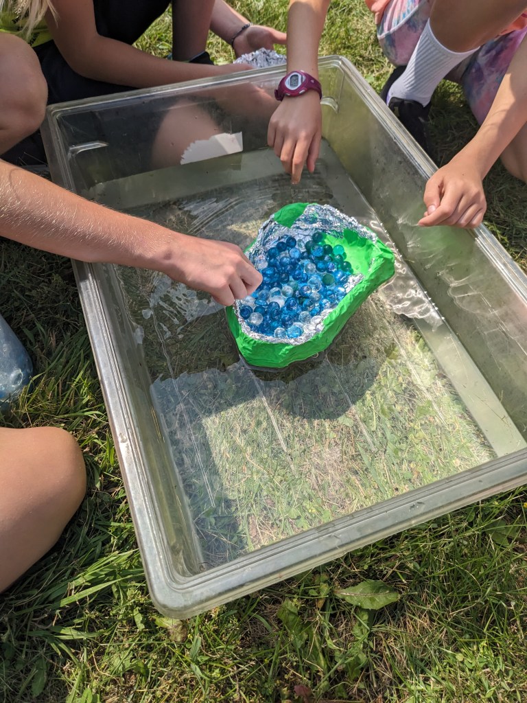 Hands placing handmade tin foil boats into a tub of water to test buoyancy.
